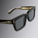A close up image of Black sunglasses by Cutler and Gross with gold accents on a gray background
