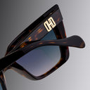 An image of the Cutler and Gross Valentina 002 Cat Eye Sunglasses in tortoiseshell frame detail close up of right hinge and temple with gold logo in grey background.