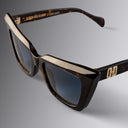 An image of the Cutler and Gross Valentina 002 Cat Eye Sunglasses in tortoiseshell frame detail close up of left lens and right temple with gold logo in grey background.