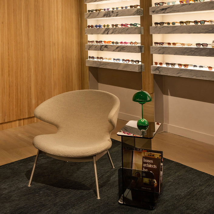 Image of inside the New York Store. In the centre of the image there is a beige chair and glass table a green lamp on. The shelves in the background have sunglasses and opticals on.