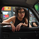 A woman with curly hair wearing the Cutler and Gross 9347 in black leans out of a car window at night, wearing a black sleeveless top. The background shows blurred city lights.