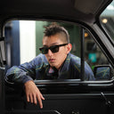 A man wearing the Cutler and Gross 1434 black rectangular sunglasses and a blue patterned jacket leans out of a car window, looking directly at the camera.