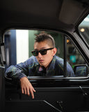 A man with a mohawk hairstyle wearing black sunglasses and a blue plaid shirt leans out of a car window, looking directly at the camera.
