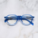 An image of the GR06 Round Optical Glasses in Blue Crystal frame frontal view with arms closed marble background 